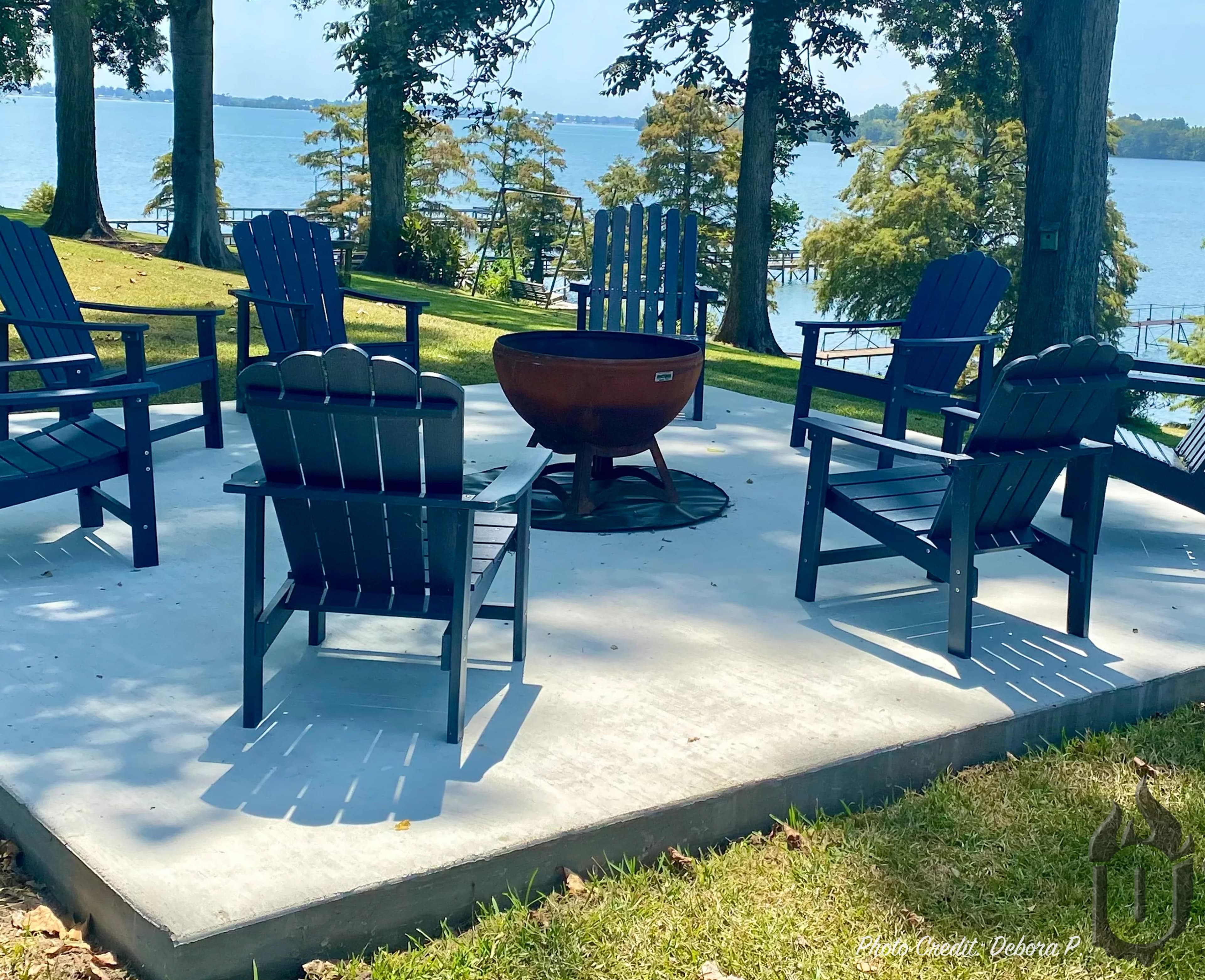 Ohio Flame 37-inch Fire Chalice artisan fire bowl with natural rust patina installed on a lakeside concrete patio surrounded by six navy Adirondack chairs, mature oaks and a wide open lake view in the background.