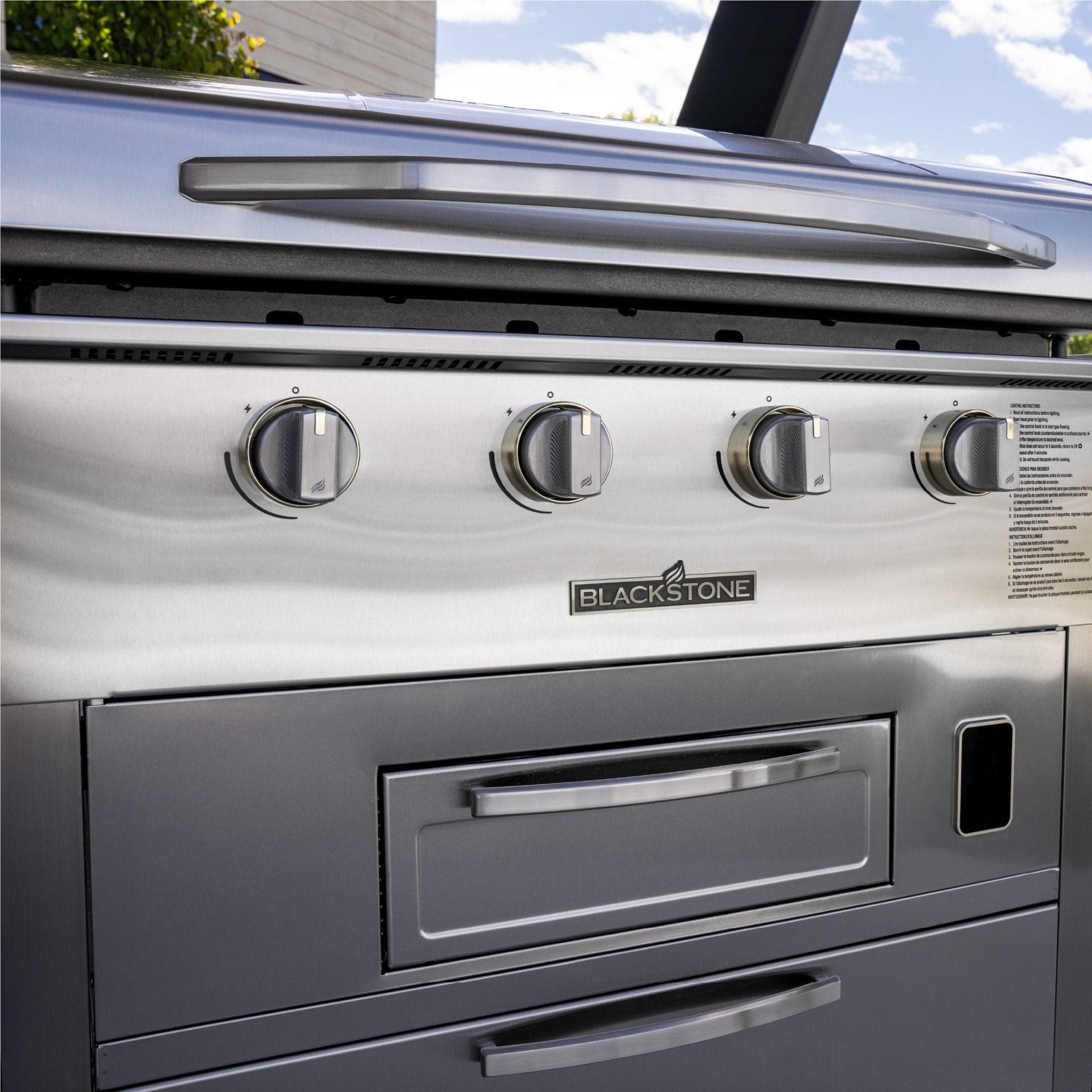 Close-up of the Blackstone Select 36-inch Air Fryer Combo control panel against a blue sky, showing four burner knobs, the Blackstone badge, and the stainless cabinet with soft-close drawer.
