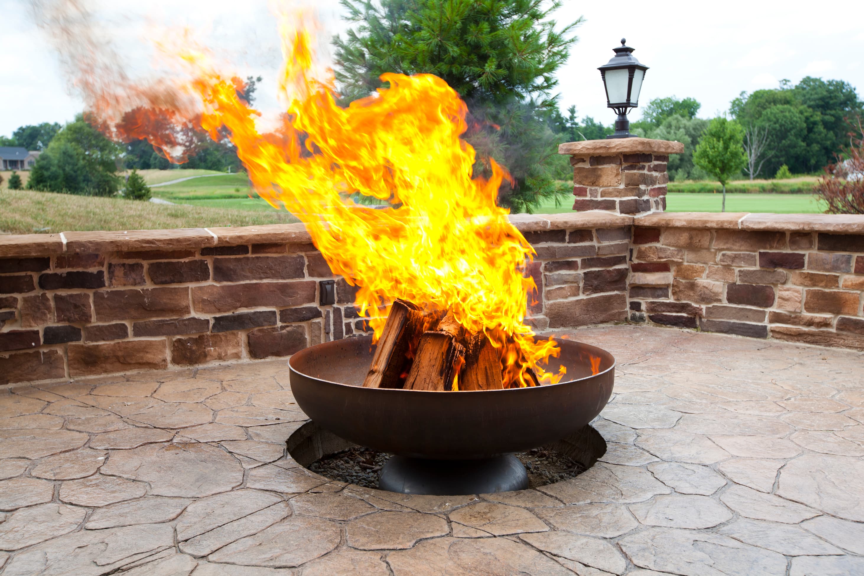 Ohio Flame Patriot 36-inch wood-burning fire pit in Corten natural steel finish, on a paver patio at dusk with four chairs pulled around it.