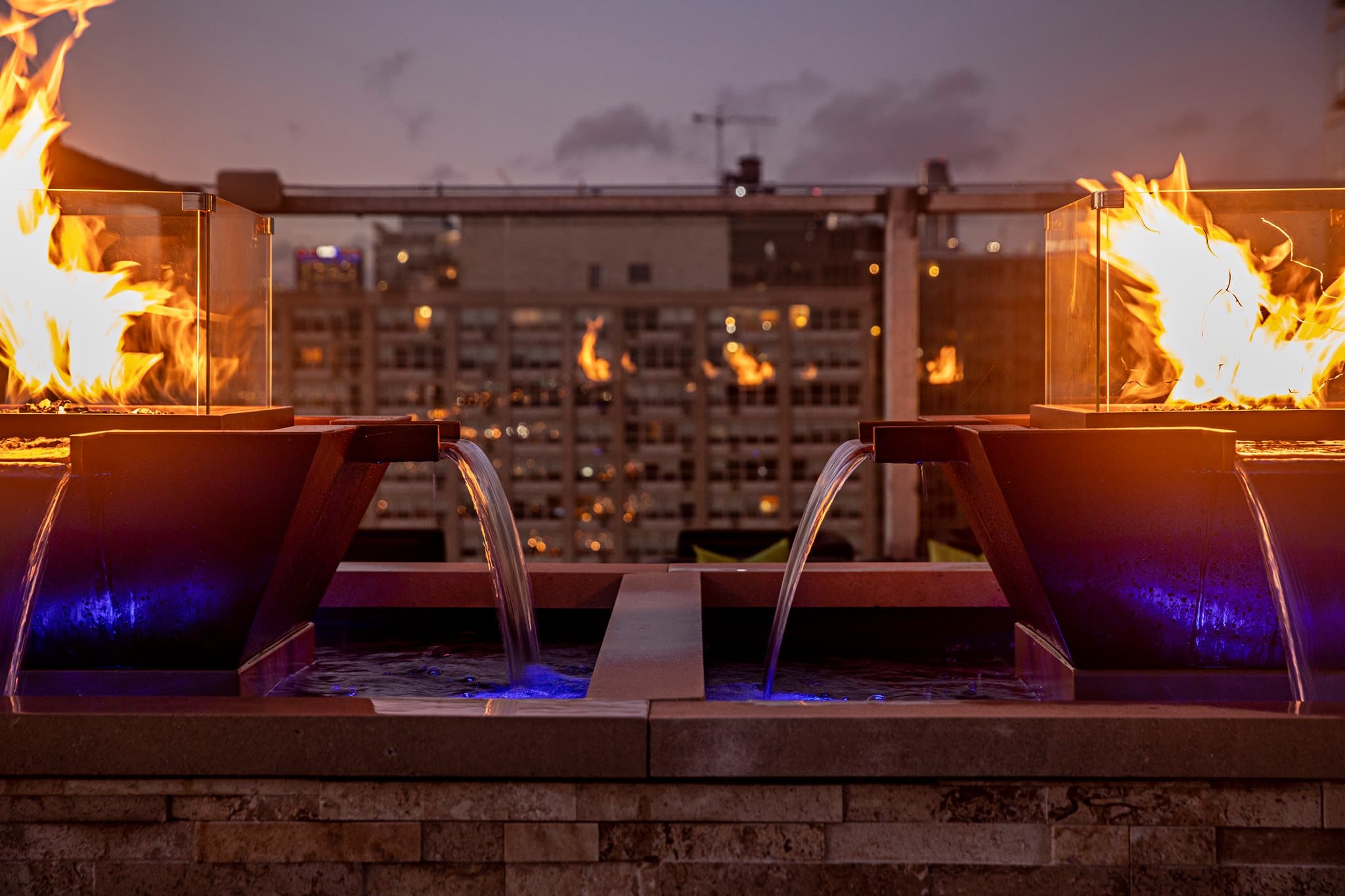 The Outdoor Plus Maya 4-way hammered copper fire and water bowl installed at the corner of a modern pool deck, flames and water flowing simultaneously — twilight patio scene.